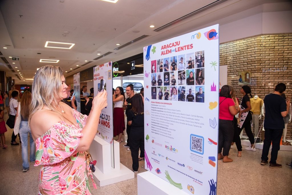 Mulher loira segura celular e fotografa um dos totens da exposição Aracaju Além das Lentes