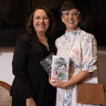 Fotografia de duas mulheres de pé, lado a lado, sorrindo para a câmera em um ambiente interno. À esquerda, uma mulher de pele clara, cabelos castanhos ondulados na altura dos ombros, usa óculos de grau com armação dourada, colar de pérolas, blusa preta e um blazer preto. Ela segura um livro com capa escura parcialmente visível. À direita, uma mulher de pele clara, cabelos curtos e escuros, usa óculos de grau com armação clara e transparente. Ela veste um vestido branco com estampa de folhas azul-esverdeadas e flores rosadas, que possui mangas três quartos franzidas e gola alta. Ela segura, com as duas mãos, um exemplar do livro "Sandra - Caminhando", que mostra na capa a ilustração de uma criança em um caminho arborizado. Ela também carrega uma bolsa tiracolo marrom com as letras "SCH" gravadas na parte frontal. Ao fundo, observa-se uma parede escura e parte de uma cadeira com encosto de palha à esquerda. A iluminação é suave e focalizada nas pessoas.