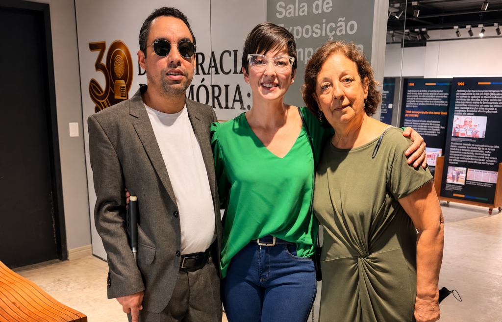Fotografia em plano médio de três pessoas em pé, lado a lado, em um espaço de exposição. Lucas, eu e Ana de Fátima, todos sorridentes para a câmera. O fundo mostra a exposição. 