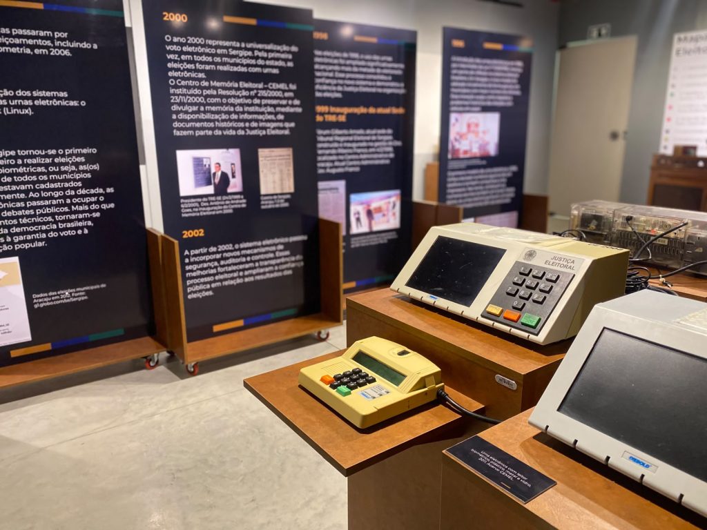 Fotografia em plano médio, focada em uma exposição museológica sobre a história das urnas eletrônicas no Brasil. No primeiro plano, à direita, três modelos diferentes de urnas eletrônicas estão dispostos sobre suportes de madeira.