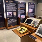 Fotografia em plano médio, focada em uma exposição museológica sobre a história das urnas eletrônicas no Brasil. No primeiro plano, à direita, três modelos diferentes de urnas eletrônicas estão dispostos sobre suportes de madeira.