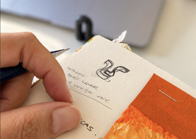 Minha mão canhota segura uma lapiseira sobre um caderno pequeno aberto. Na página, o desenho a lápis do número 25 em forma de fita e, ao lado, escrito à mão: "insight após lavar a louça kkk". Ao fundo, desfocado, um notebook aberto. Na borda do caderno, papel laranja preso por grampo (teste de textura).